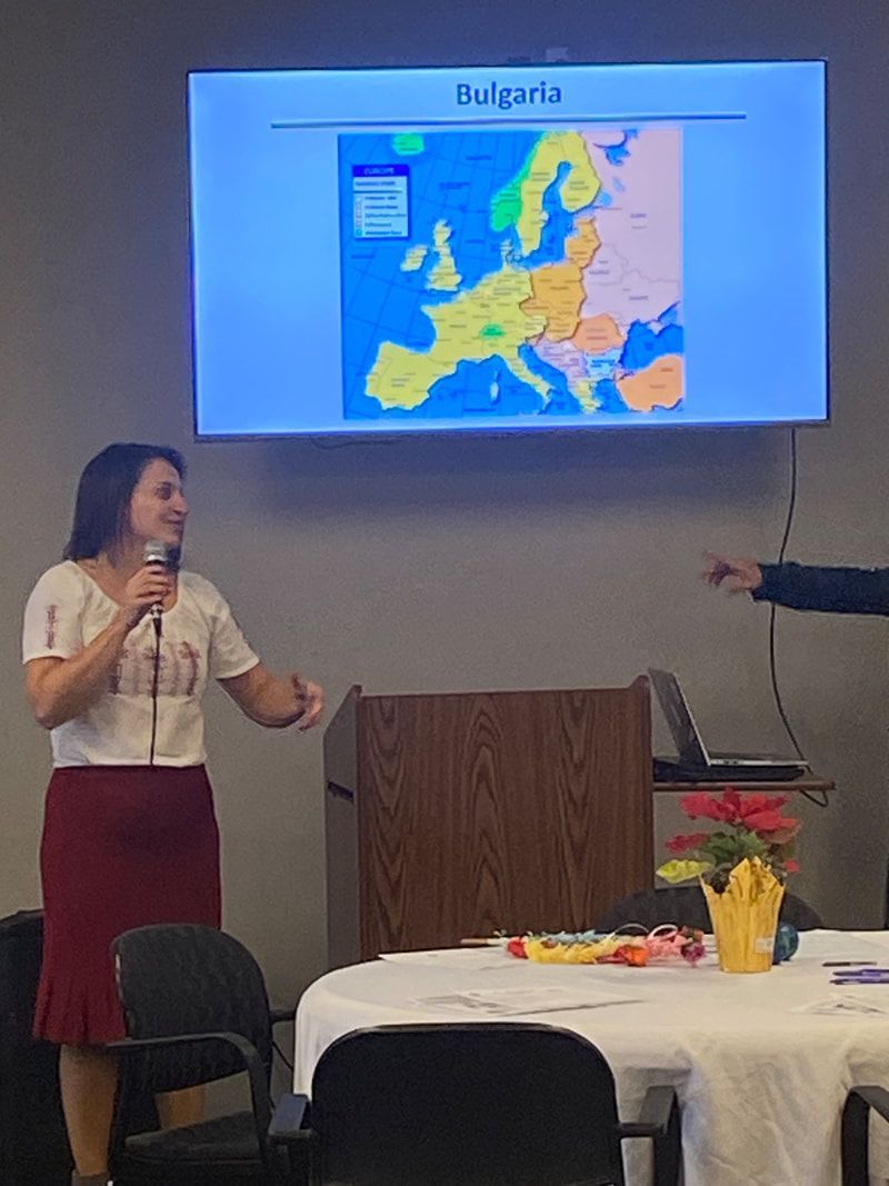 Woman presenting on Bulgaria with a map of Europe. She wears a white top and red skirt, speaking with a microphone.