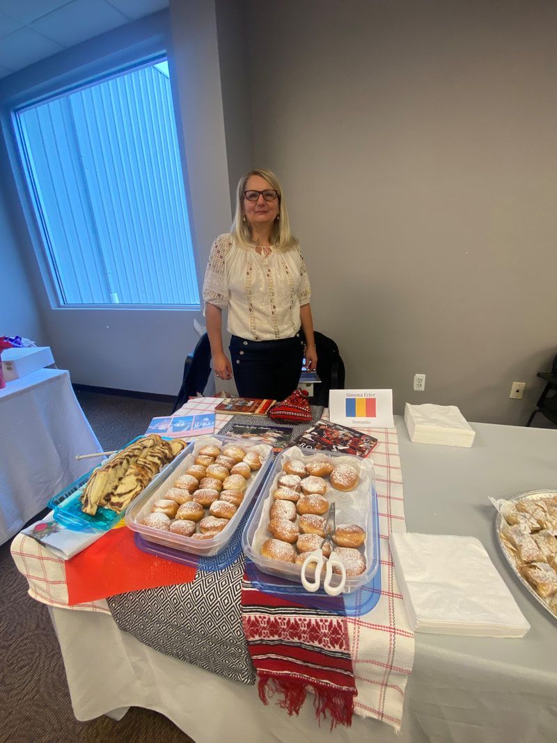 Woman stands behind a table of pastries, Romanian flag, indoors.