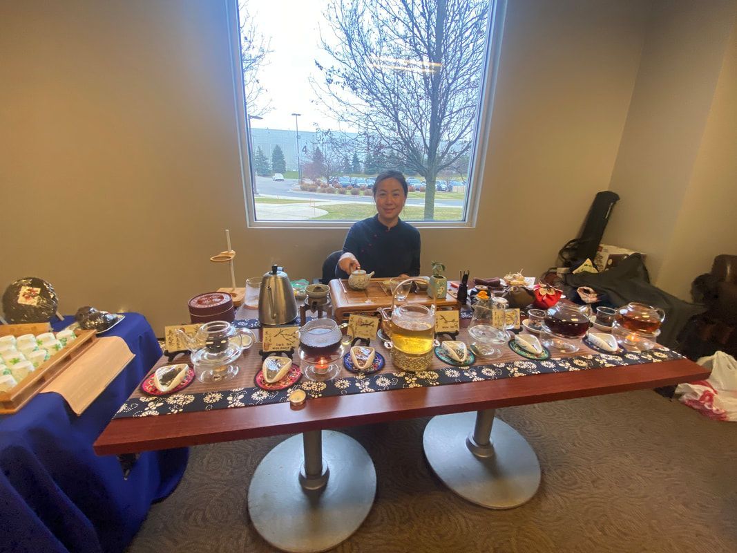 A woman serves tea at a table with various treats, set up near a window.