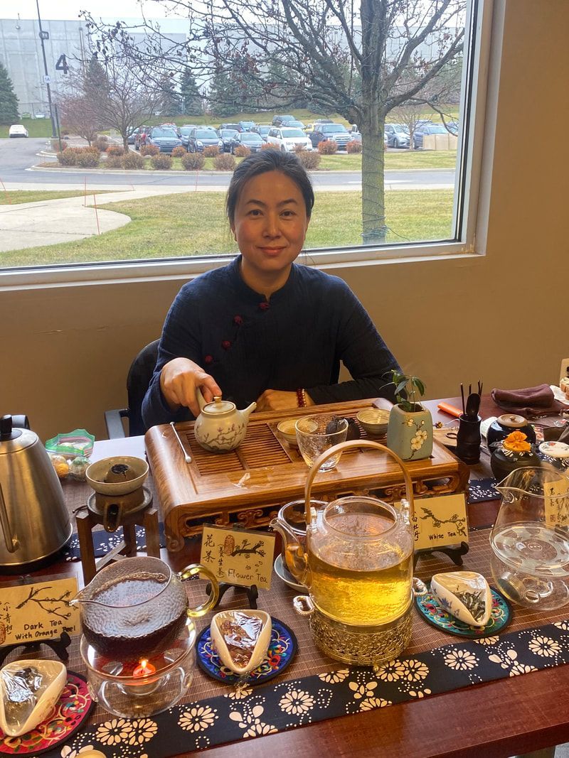 Woman pouring tea at a table set with teacups, a teapot, and tea-making accessories indoors by a window.