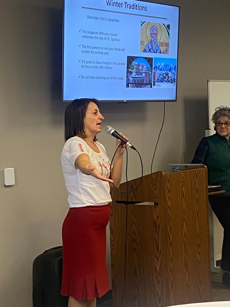 Woman in red skirt speaks at a podium, presenting slides with religious imagery. Another woman watches.