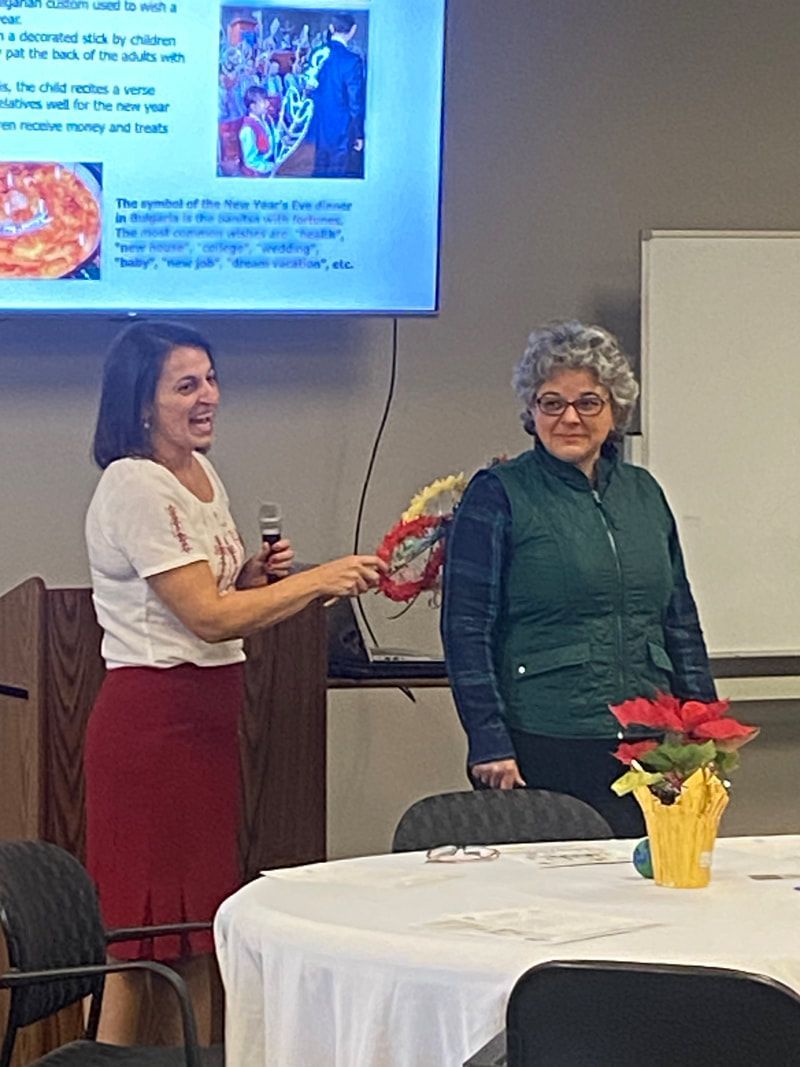 Two women at a presentation. One speaks, points with a lei. The other stands by with a smile, both by a table and projector screen.