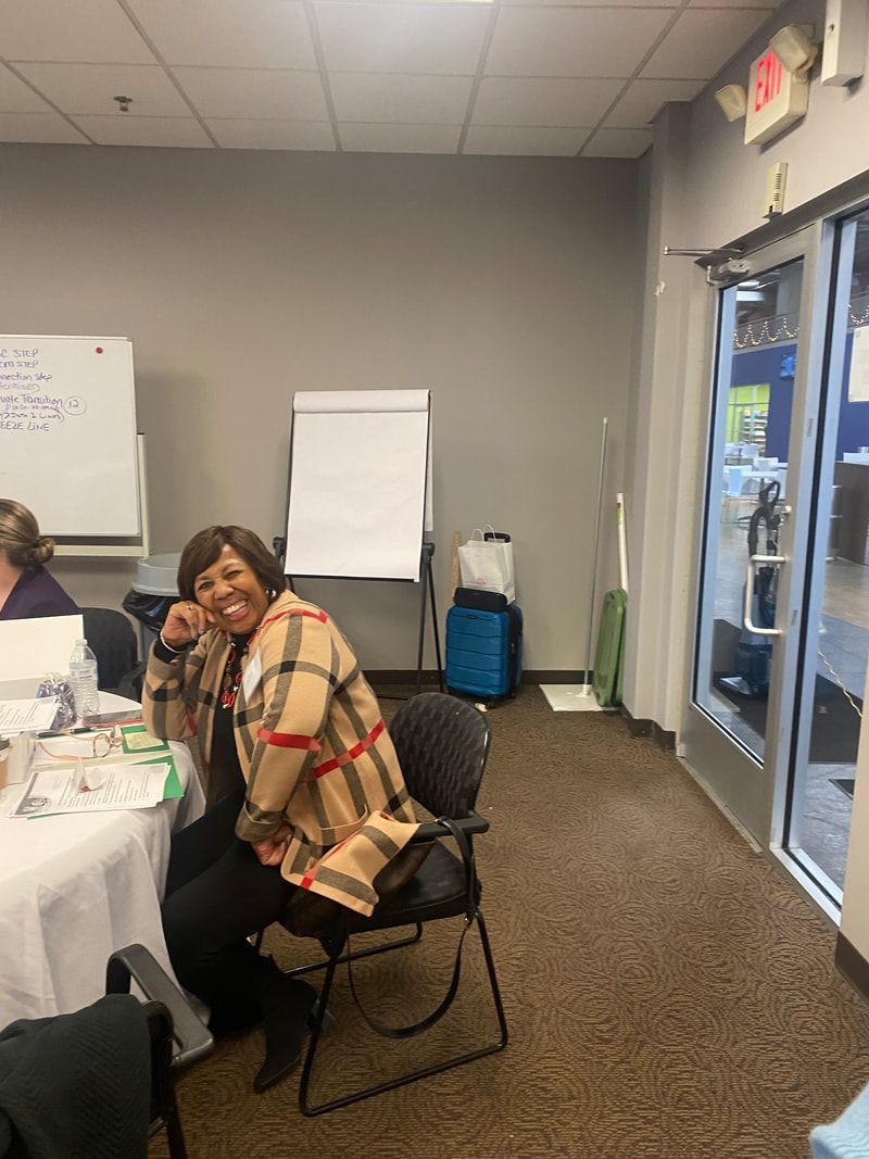 Woman in tan plaid jacket smiles in an office setting, near a flip chart and glass door.