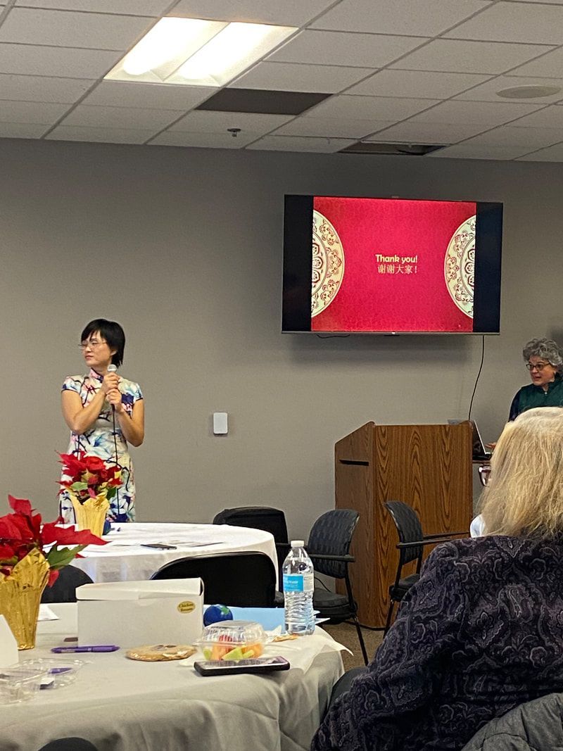 Woman in floral dress gives presentation; a screen displays a red book cover with gold trim.