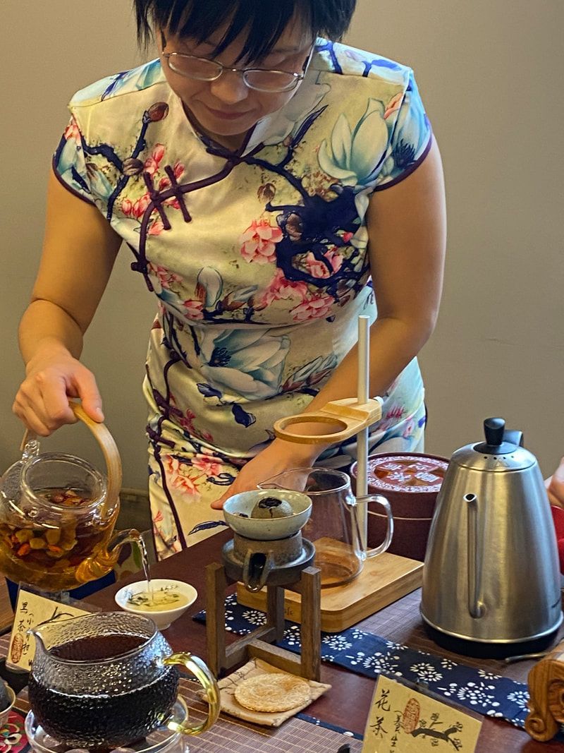 Woman in floral dress prepares tea, pouring from glass teapot. Tea ceremony setting.