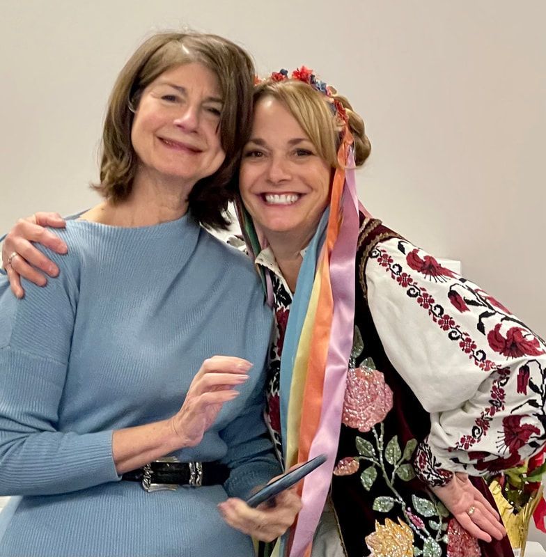 Two women pose smiling; one in Ukrainian folk costume, the other in blue.