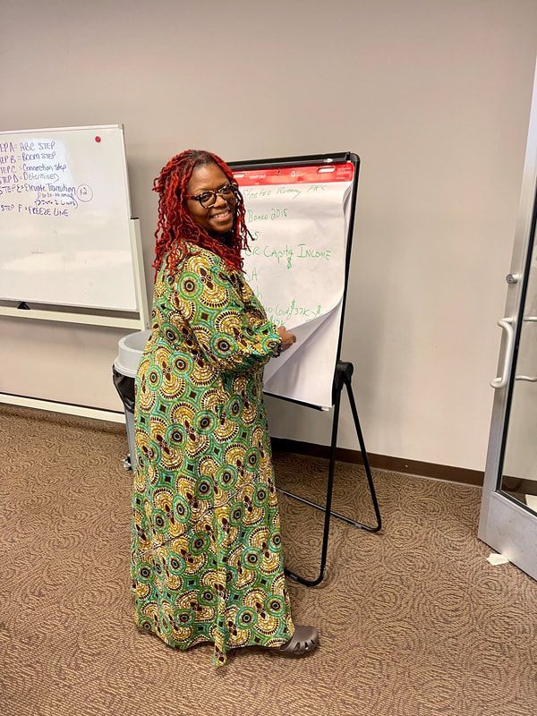 Woman in a green and yellow patterned dress smiles, turning a page on a whiteboard; indoor setting.