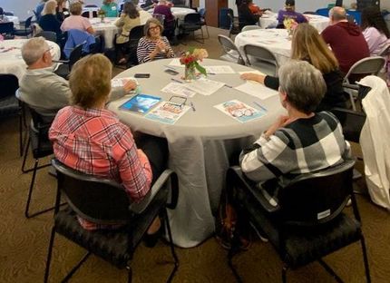 People seated at round tables in a meeting room; some are talking.