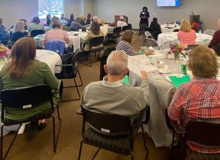 A group of people in a room with tables are listening to a woman giving a presentation.
