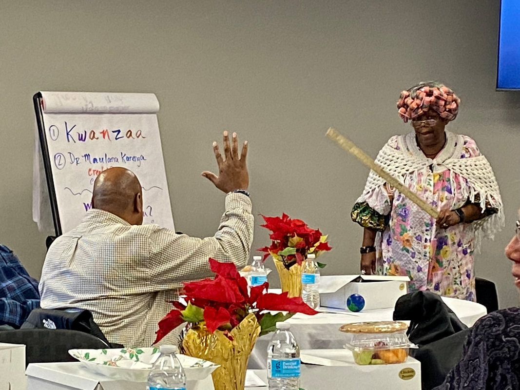 People at a Kwanzaa event. Woman with hat and stick, man raises hand. Table with flowers, easel.