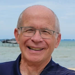 Smiling older man with glasses outdoors, blue shirt, ocean in background.
