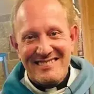 Man with fair skin, smiling widely, wearing a blue hooded garment and clerical collar.
