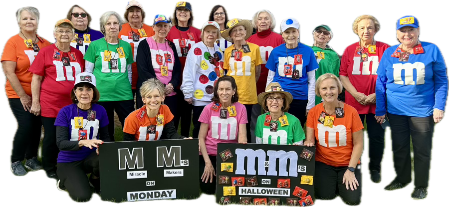 Group of people dressed as M&M candies pose with signs. Wearing colorful shirts, some hold hands, posing outdoors.
