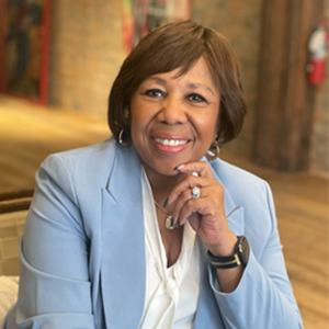 Smiling Black woman wearing a blue blazer, resting hand on her chin, indoors.