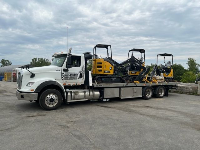 Une dépanneuse blanche avec une excavatrice jaune à l'arrière est garée sur un parking.