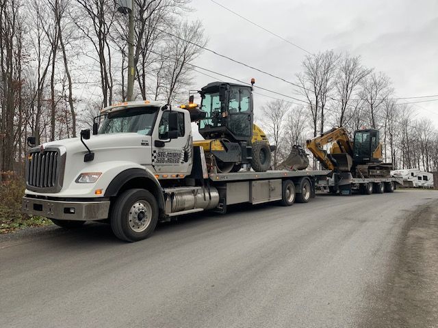 Une dépanneuse blanche transporte une excavatrice jaune sur une remorque à plateau.