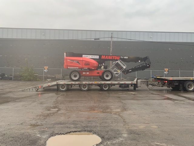 Un élévateur Manitou est posé sur une remorque à plateau dans un parking.