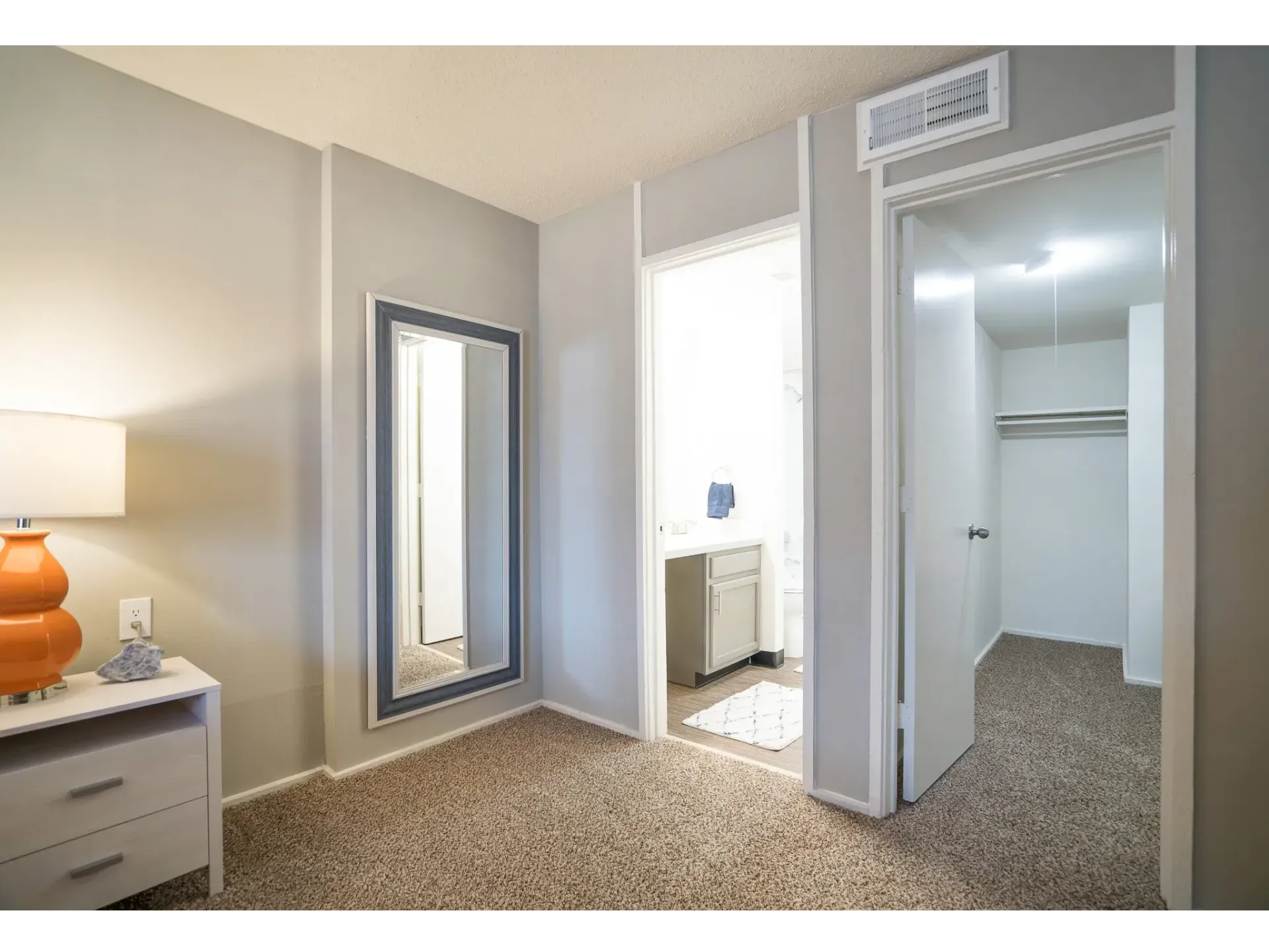A well-lit apartment interior showing a bedroom with a decorative lamp and a mirror at Haven on the Lake in Maryland Heights, MO..
