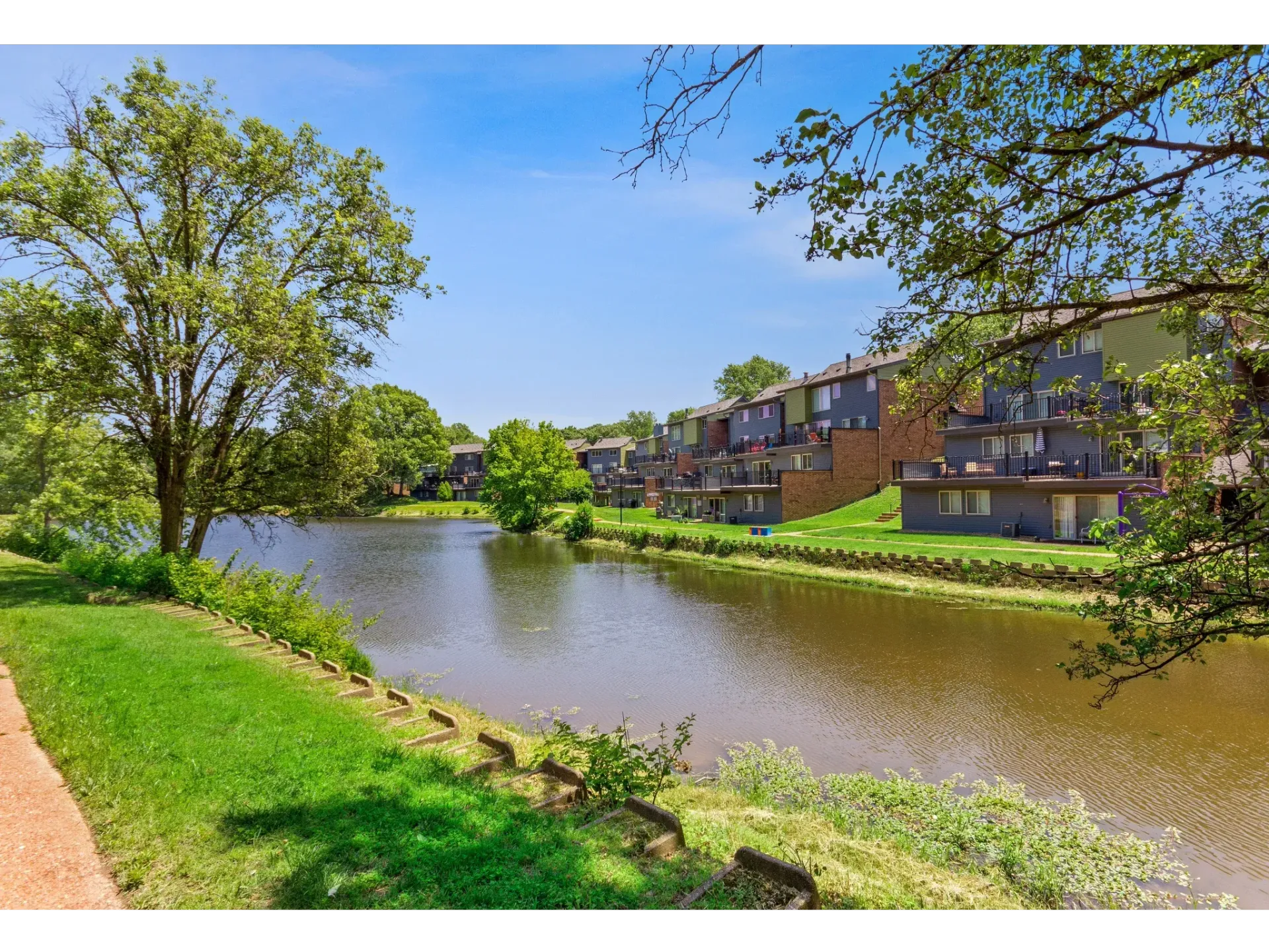 Exterior view of apartment buildings along a river with trees and a green lawn.