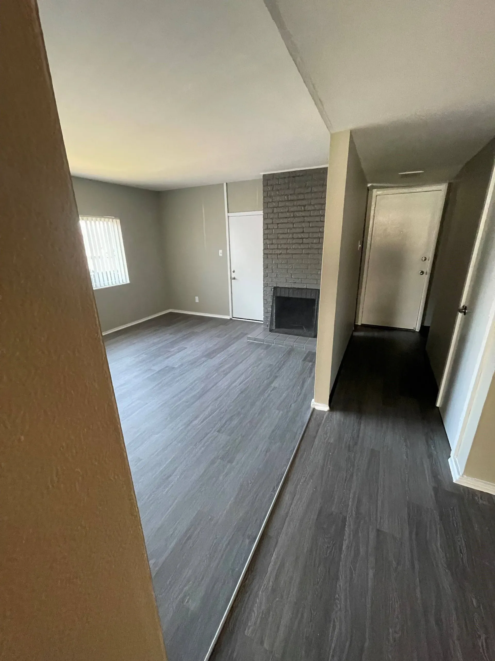 Living room with gray laminate flooring, brick fireplace, and hallway to the entry door.