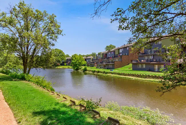 Lakeside apartment buildings under a blue sky, framed by trees and green vegetation.