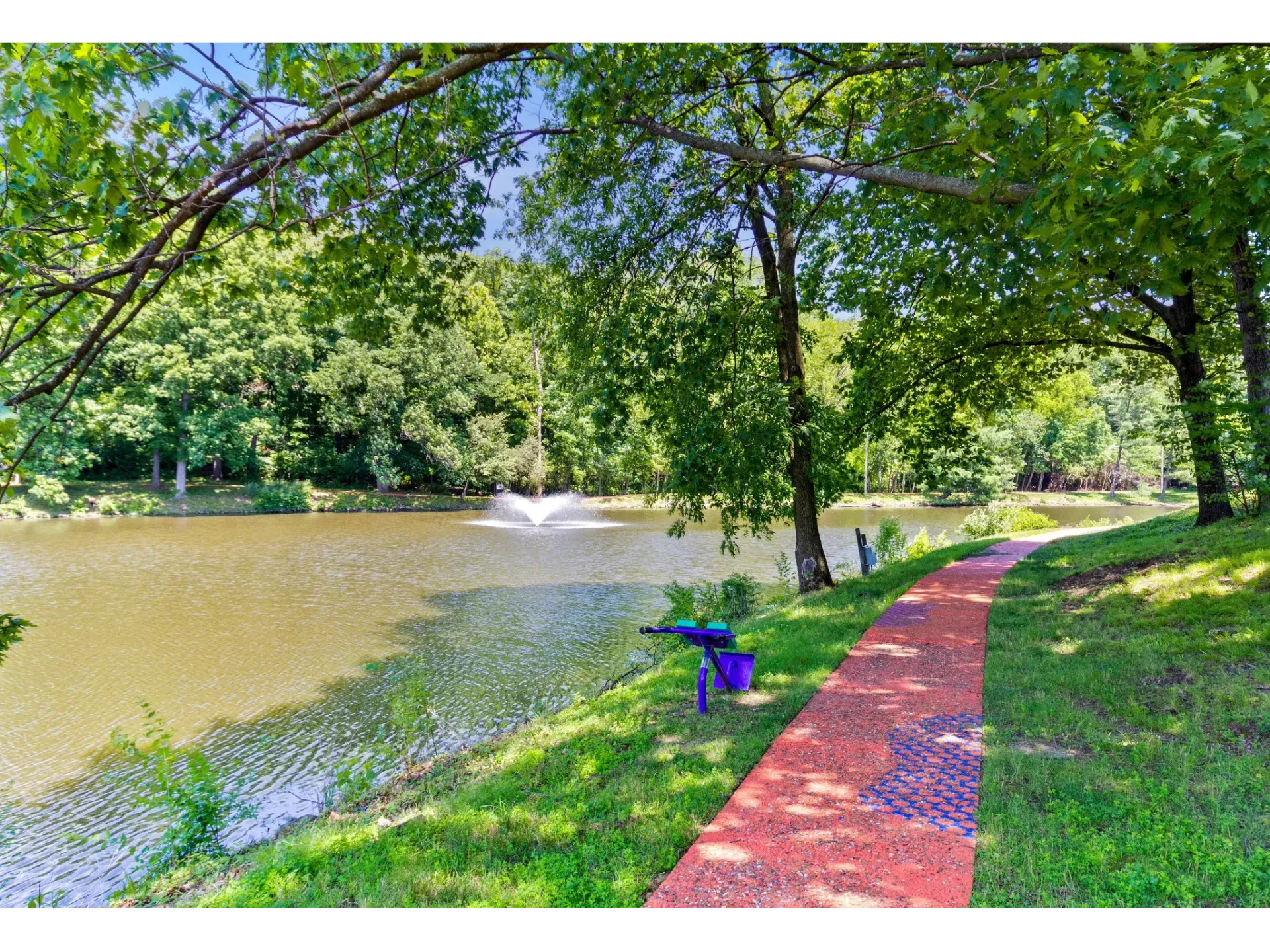 Pond with fountain, red walkway, and lush trees in a park-like apartment community.