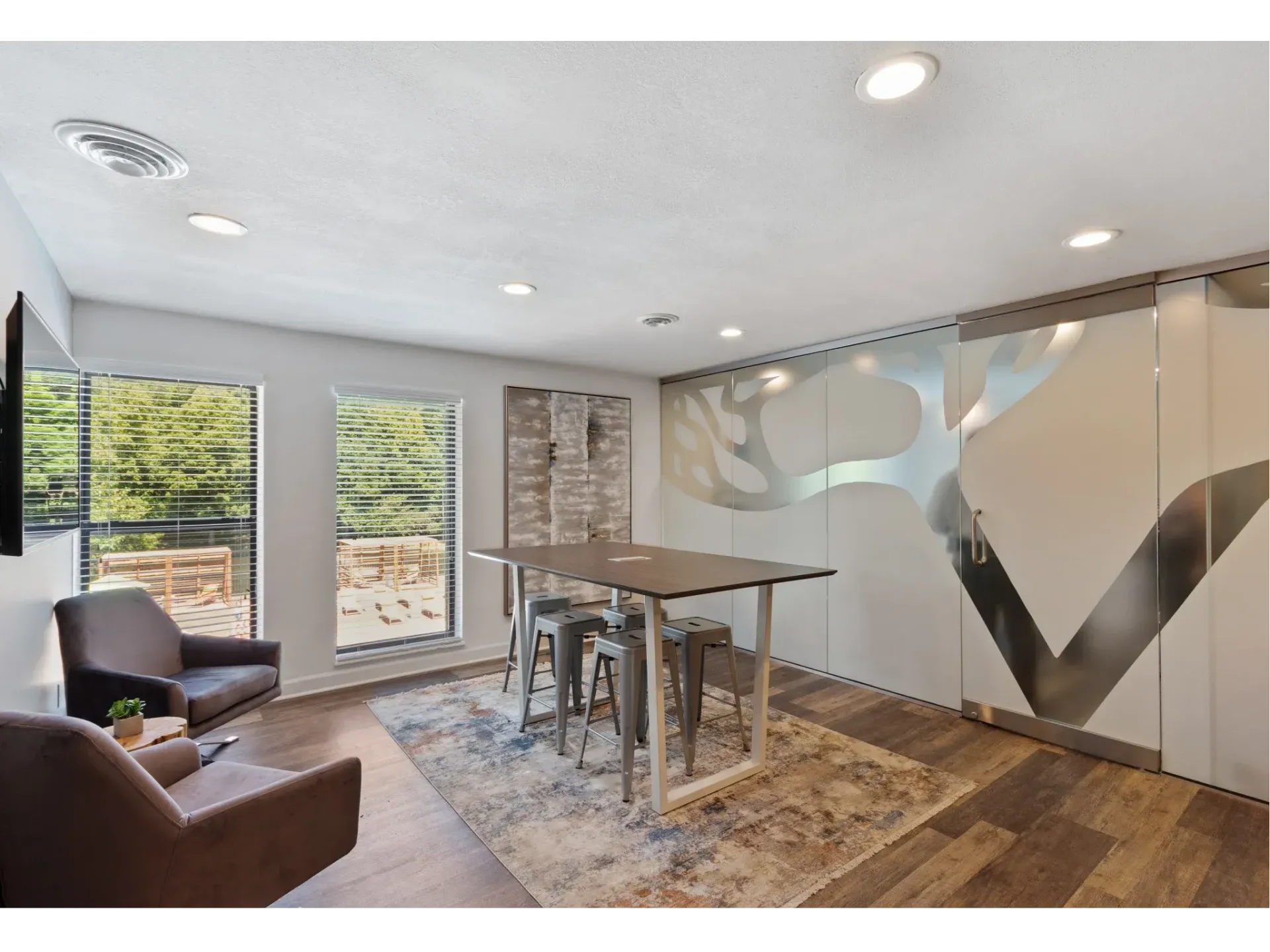 Community lounge with a high-top table, metal stools, two armchairs, large windows, and frosted glass wall.