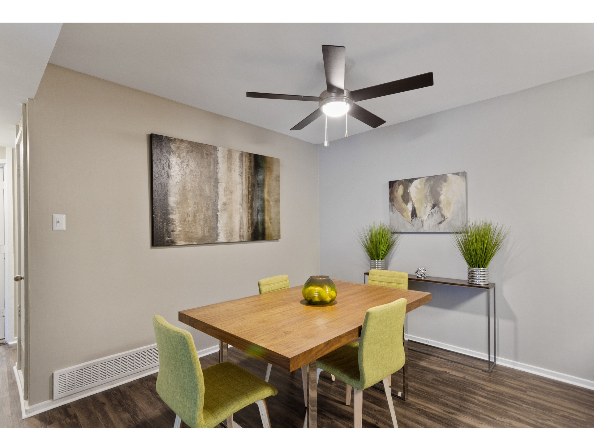 Interior dining area in an apartment with a wooden table, green chairs, a ceiling fan, and wall art.