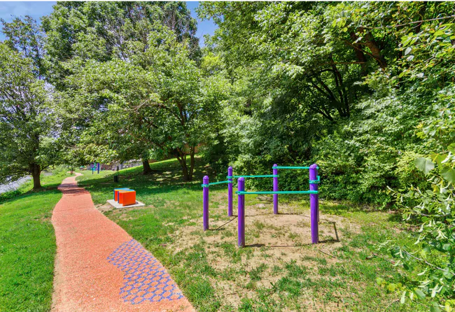Red brick path leads to outdoor exercise equipment in a grassy park, surrounded by trees.