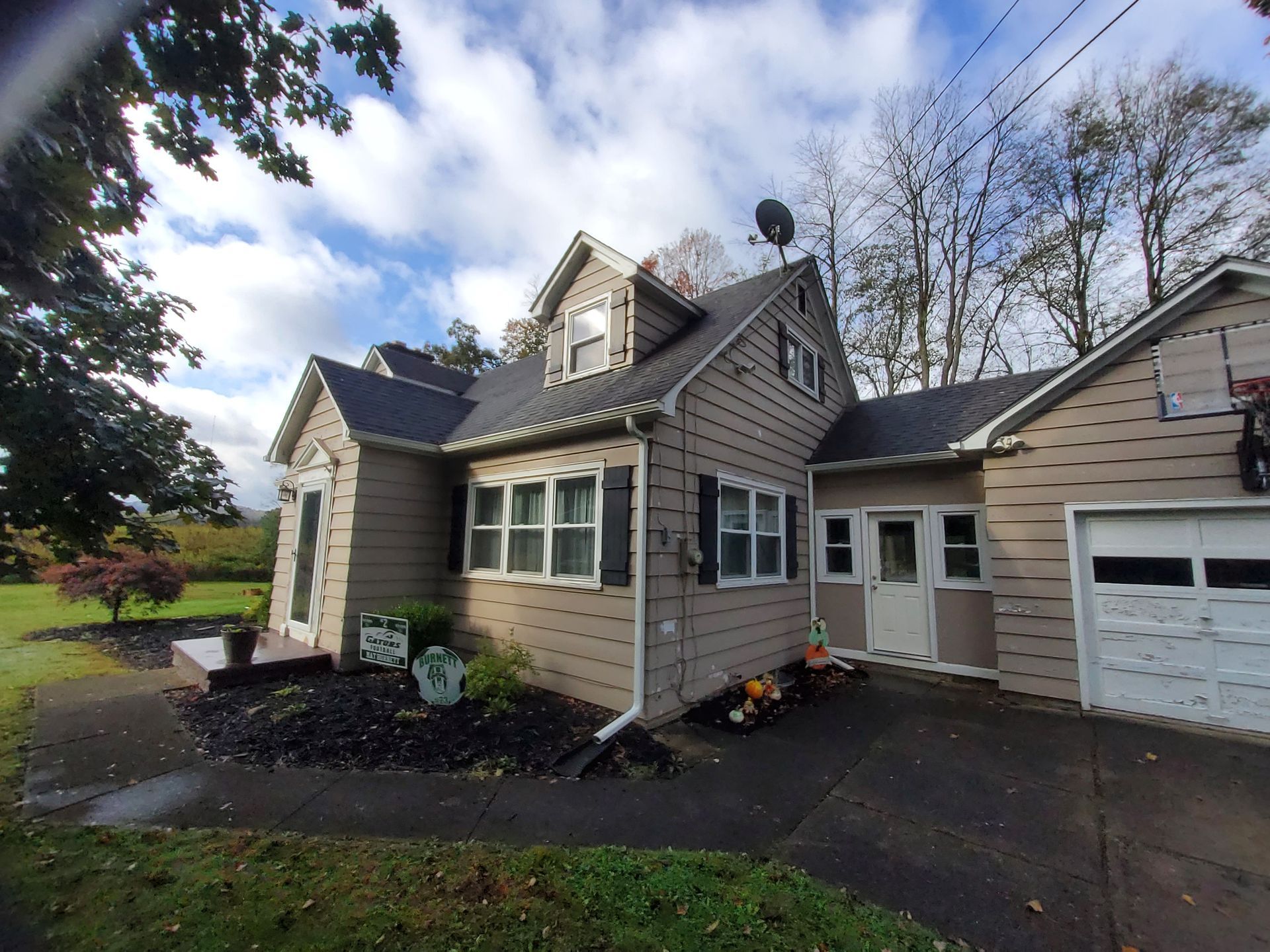 Tan two-story house with a garage, black shutters, and satellite dish against a cloudy sky.