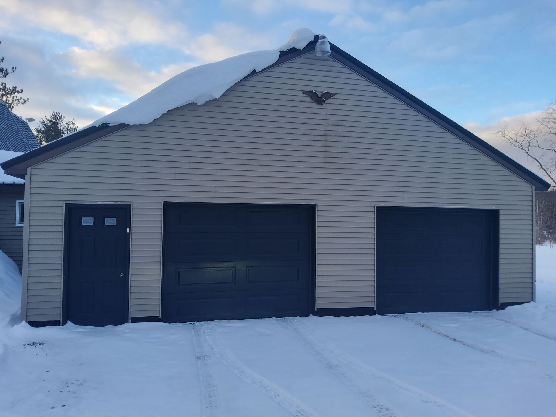 A snow-covered garage with two doors and a side door, tan siding, and a dark roof; winter scene.