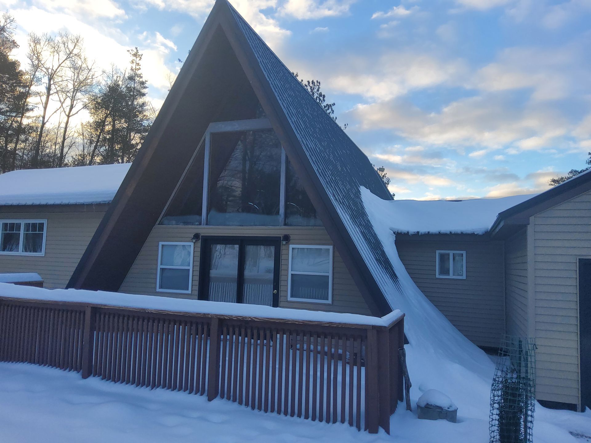 A-frame house covered in snow with a wooden deck and a cloudy sky.