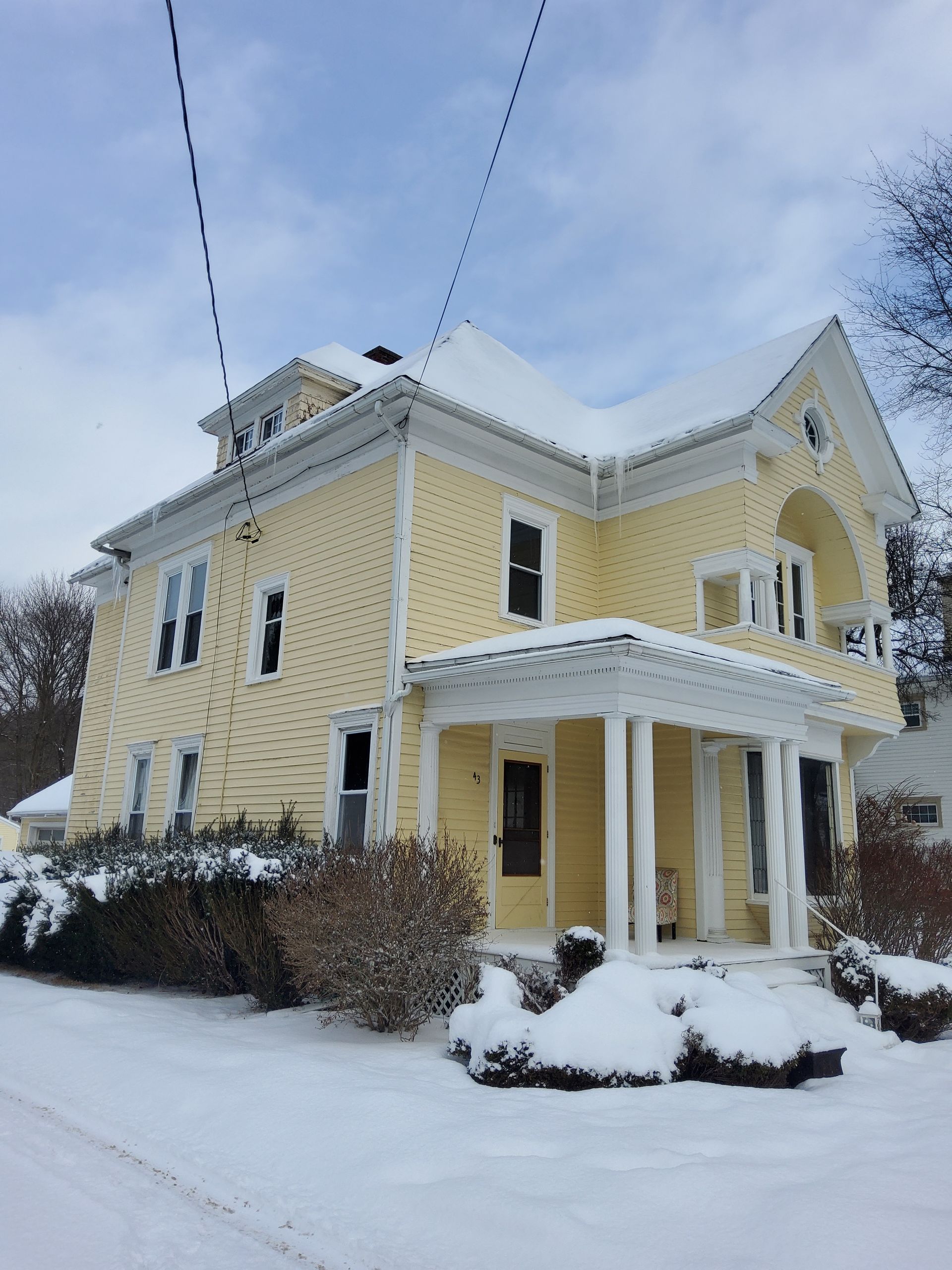 Yellow two-story house with a snow-covered roof and porch, bushes in front, and utility lines overhead.