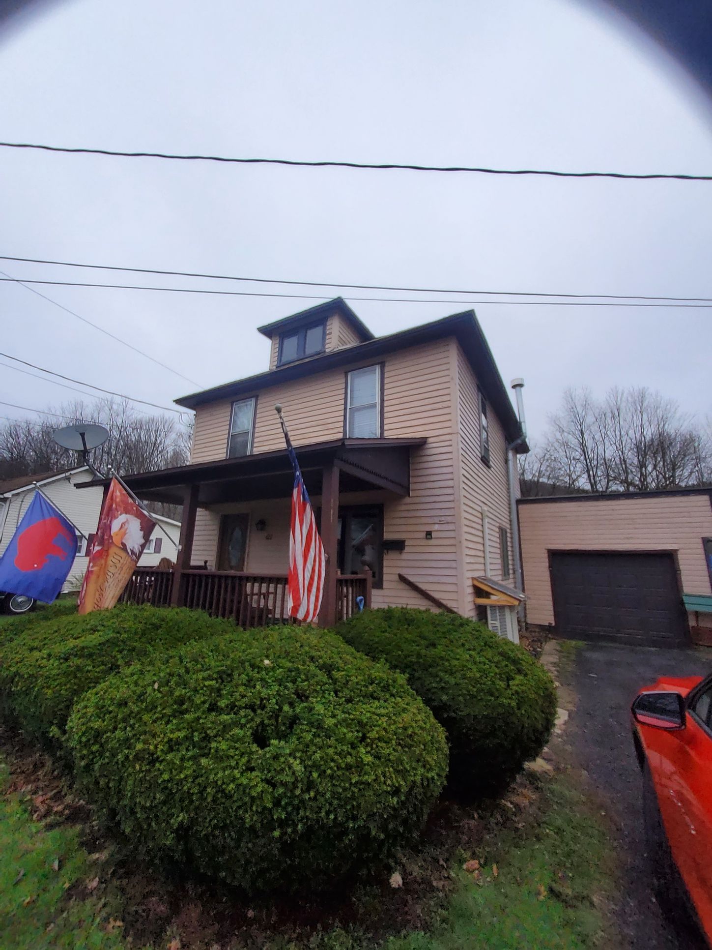 Two-story beige house with a porch and American flag. Green bushes in front; a red car to the right. Overcast sky.