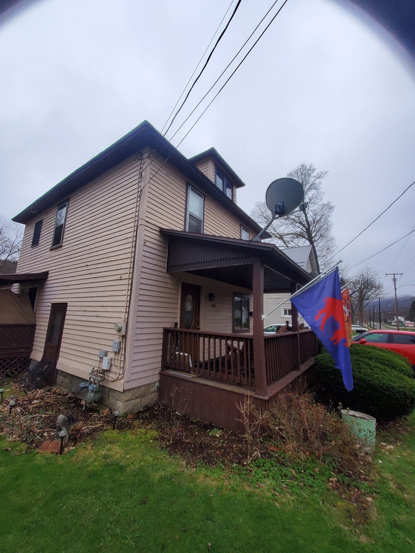 Two-story house with a porch and satellite dish, overcast sky. A flag with red design hangs on the porch.