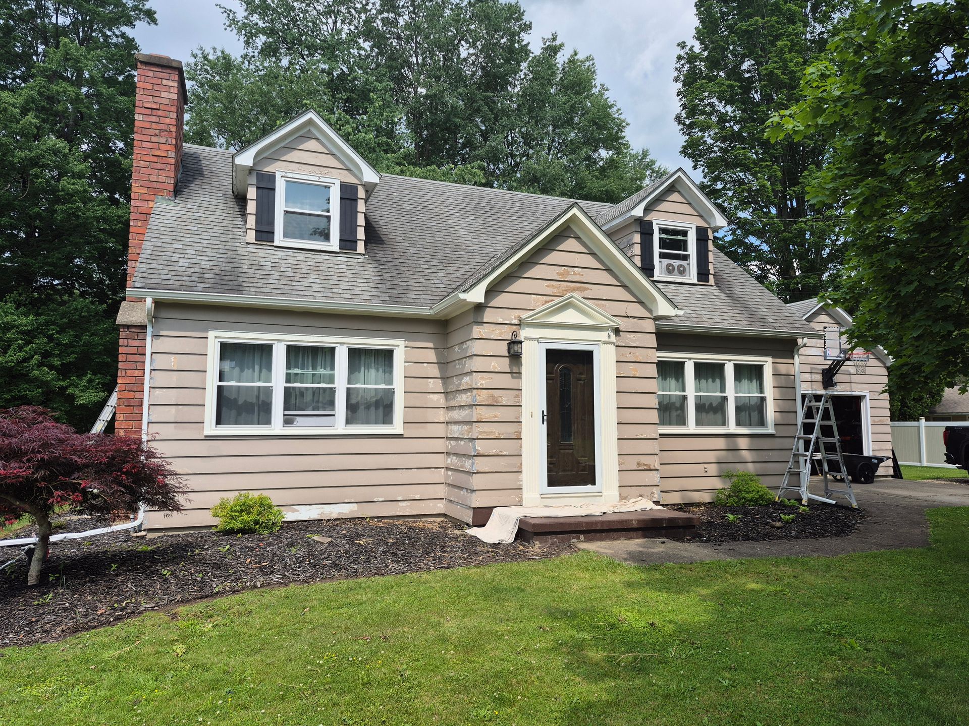 Tan house with gray roof, brick chimney, dormers. Green lawn, black mulch, trees.