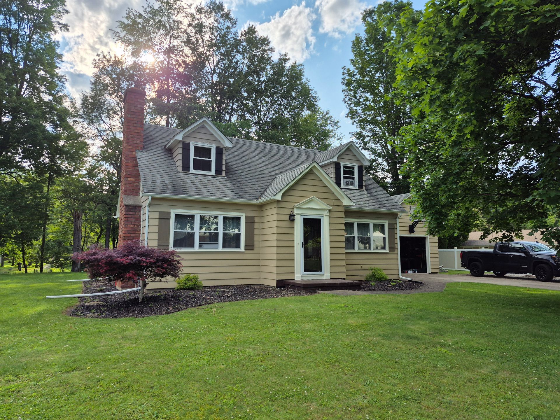 Tan house with a red brick chimney, dormers, and a garage, on a green lawn with trees.