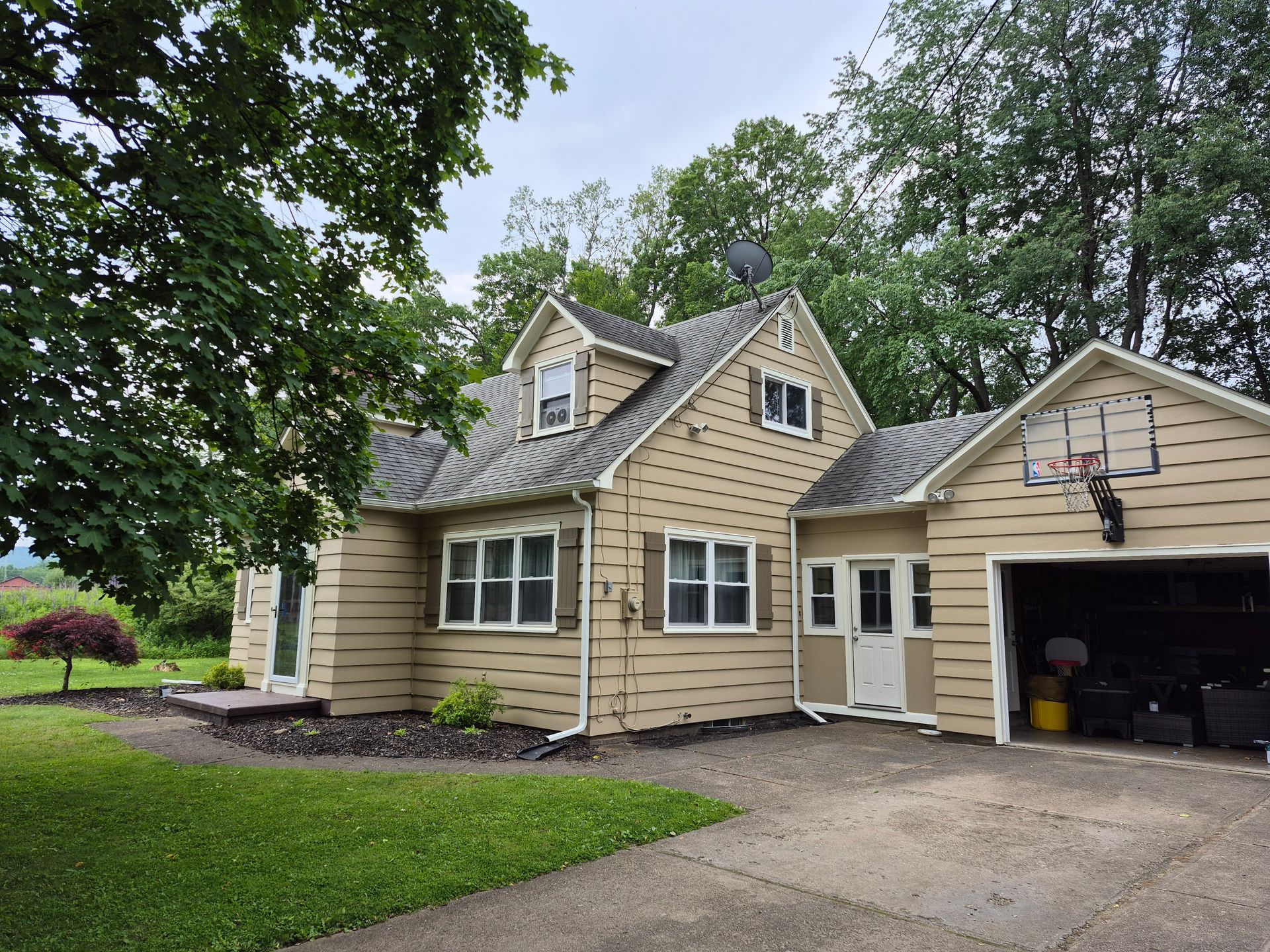 Tan house with a garage and basketball hoop. Trees surround the building; overcast sky.
