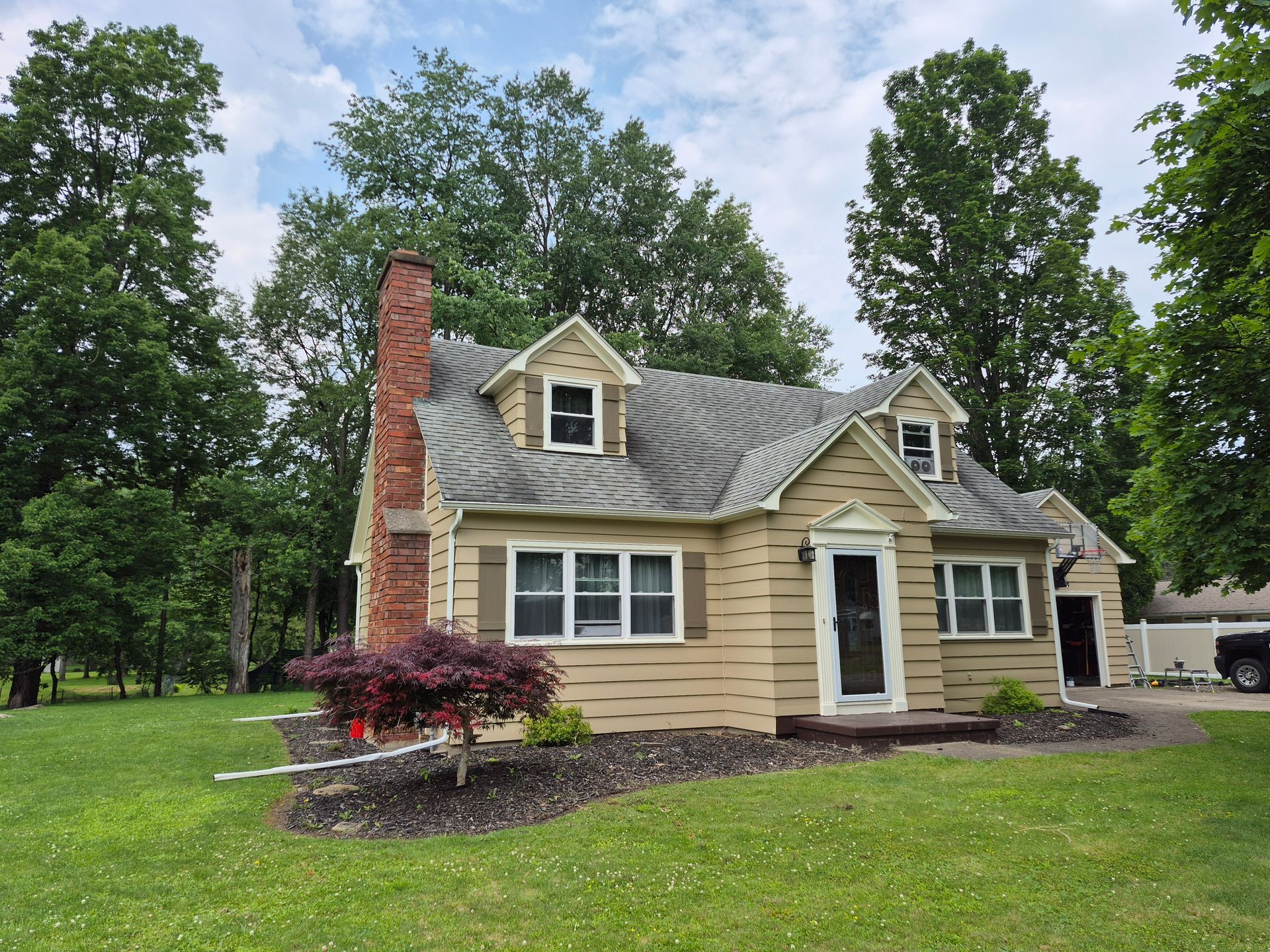 Beige house with gray roof, brick chimney, small front yard, trees, and cloudy sky.