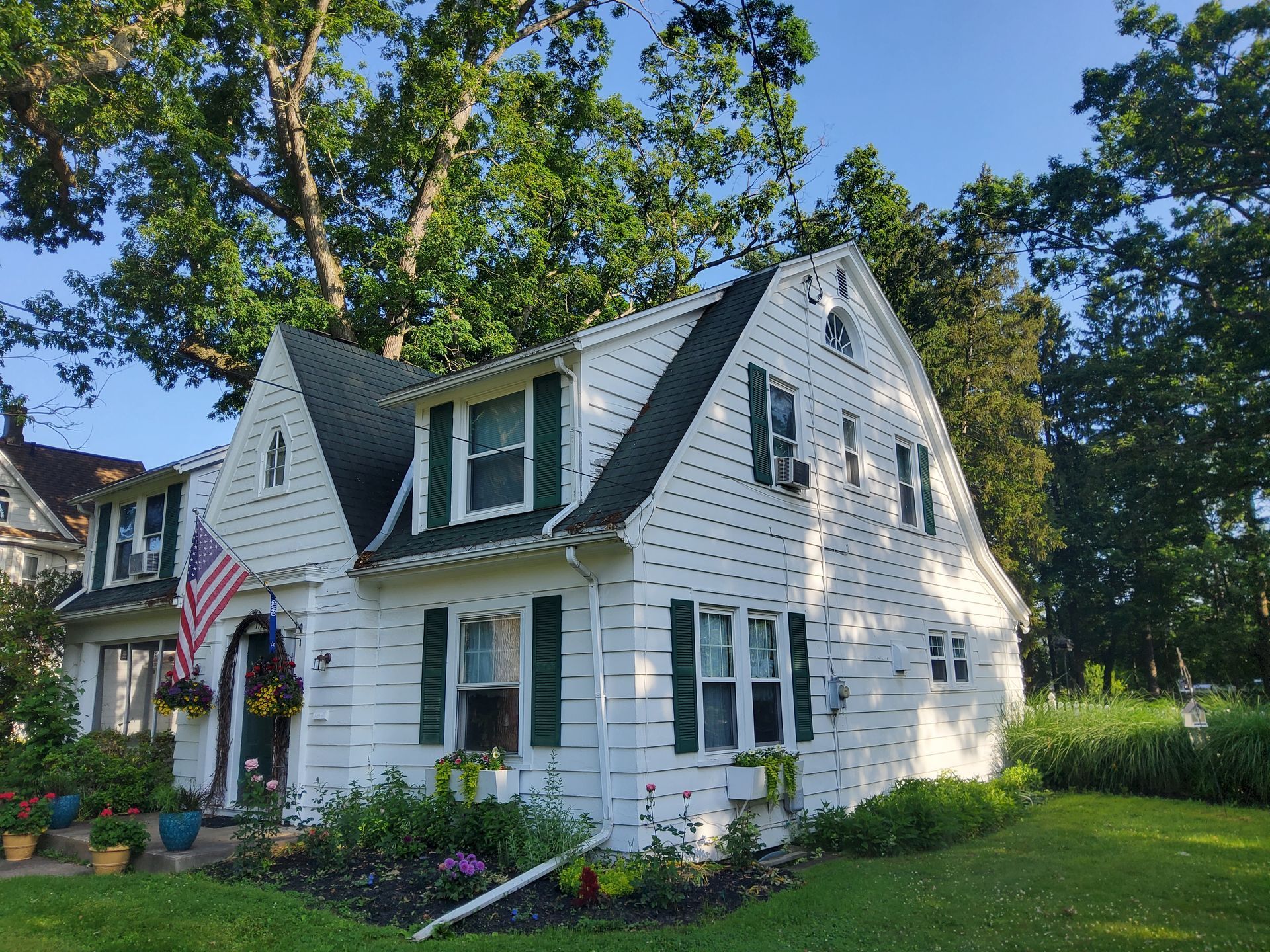 White house with green shutters, American flag, flowers, and trees in front.