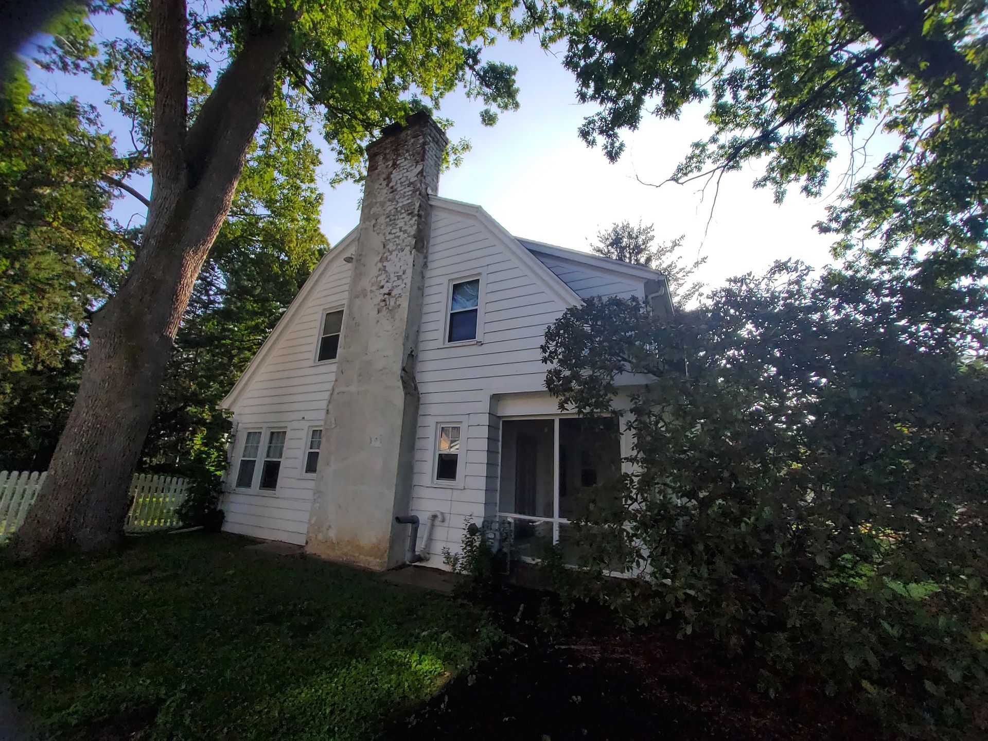 White two-story house with a large chimney. The house is surrounded by green trees and grass under a bright sky.