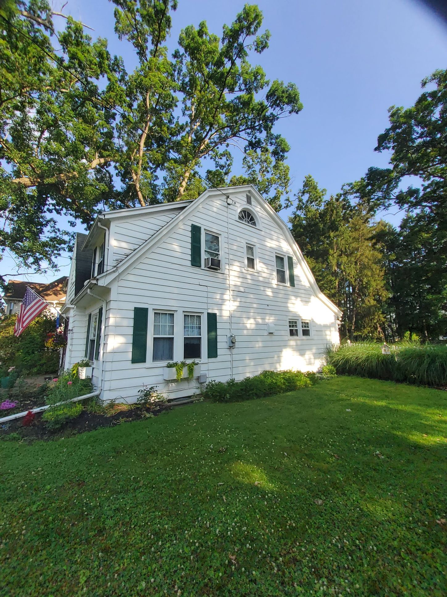 White house with green shutters, nestled amongst green trees and grass, under a blue sky.