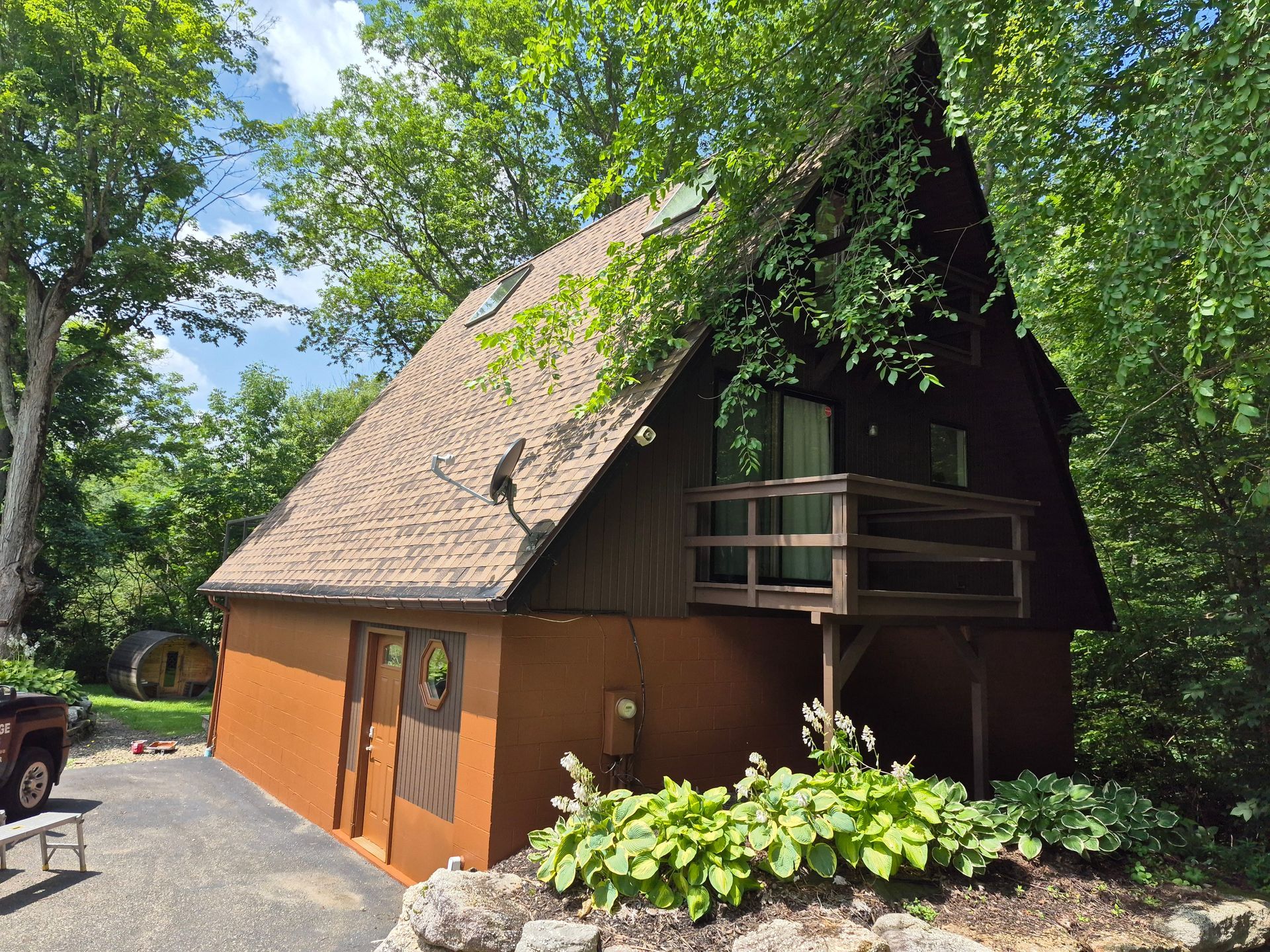 Brown A-frame house with a balcony, surrounded by trees and greenery.