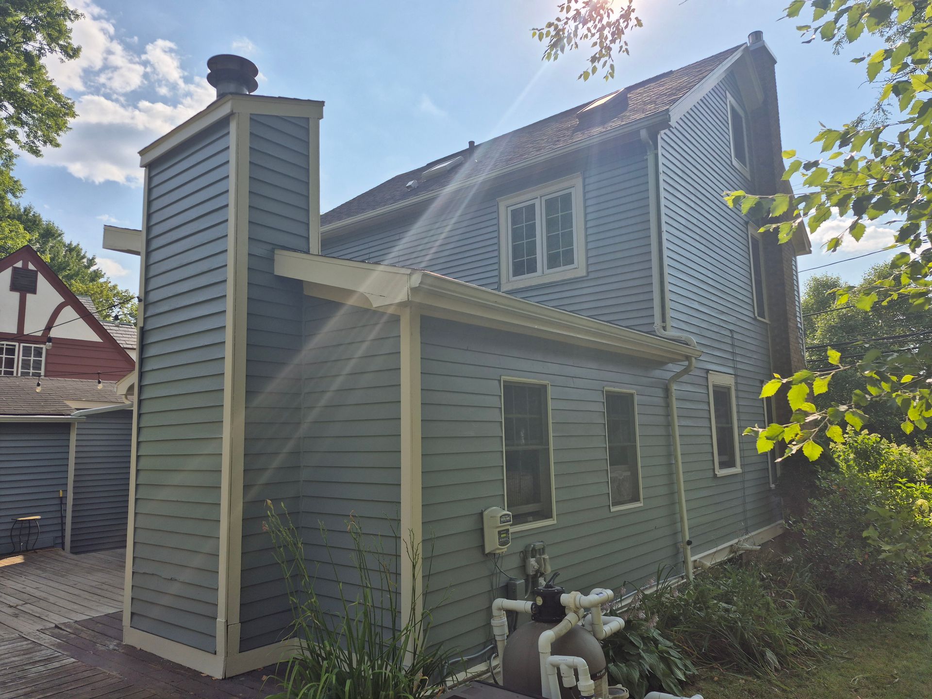 Blue-sided house with a tall chimney. White-trimmed windows and a partial view of another house with red trim are visible.