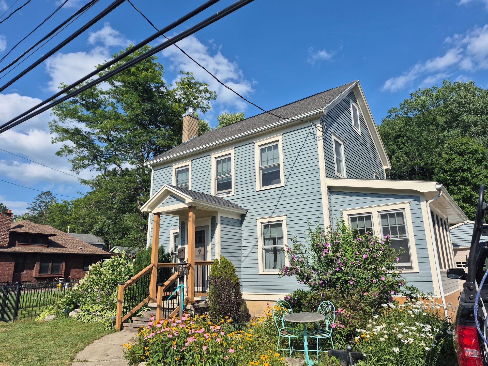 Blue two-story house with a porch and flower garden under a blue sky, trees in the background.