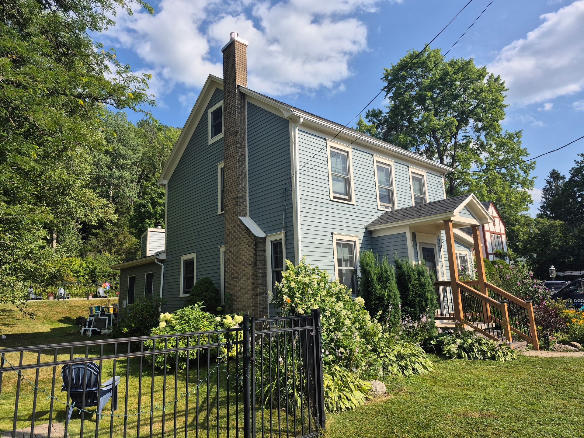 Blue two-story house with black fence, chimney, and lush greenery. Sunny day.