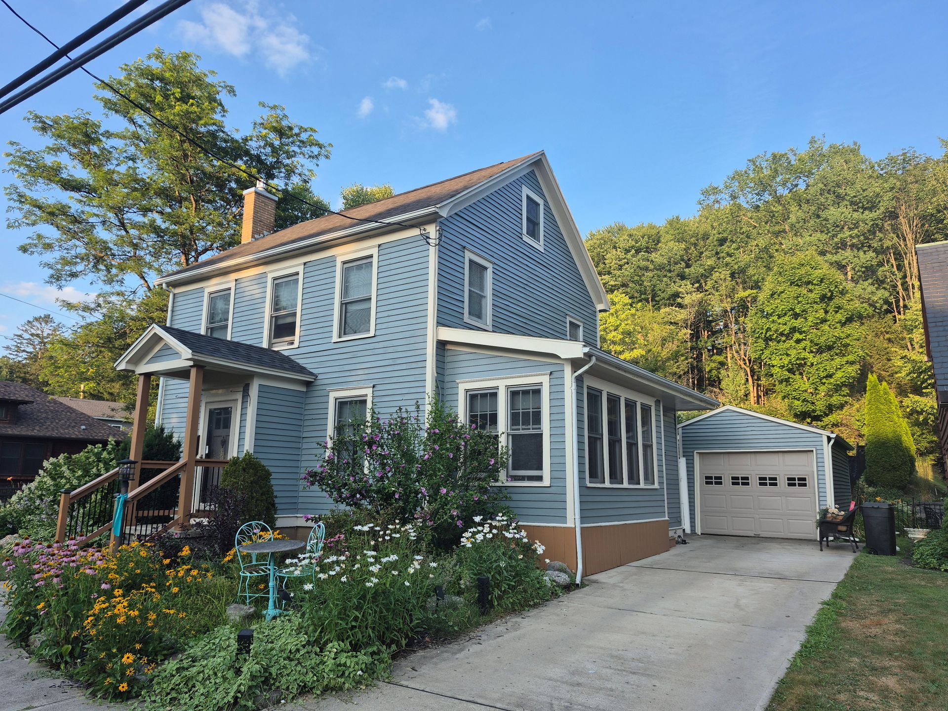 Blue two-story house with a porch and attached garage; flowers in front yard, trees in the background.