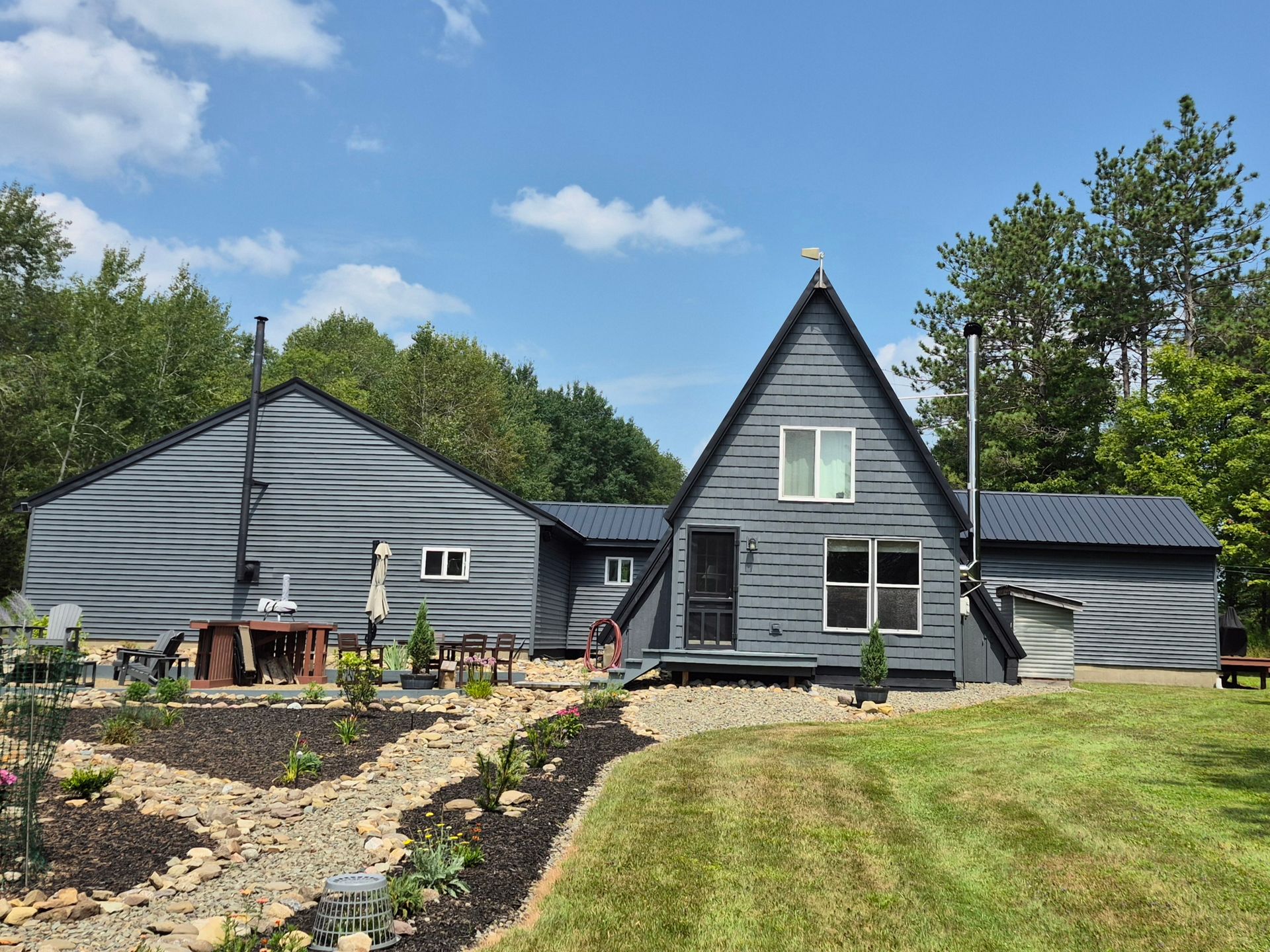Gray A-frame house with attached wing, green lawn, garden, and blue sky.