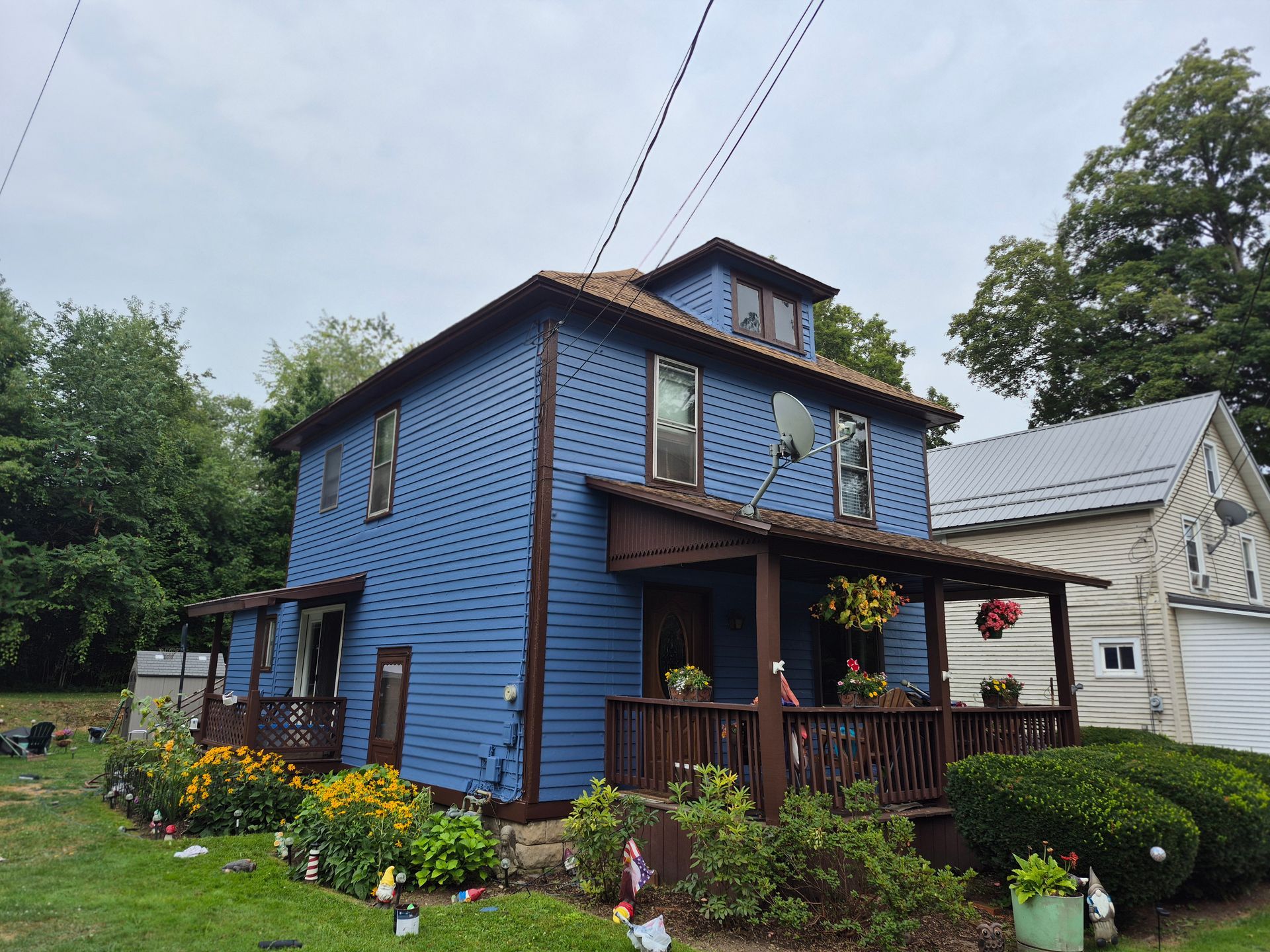 Blue two-story house with brown trim and porch, surrounded by greenery and flowers under a cloudy sky.