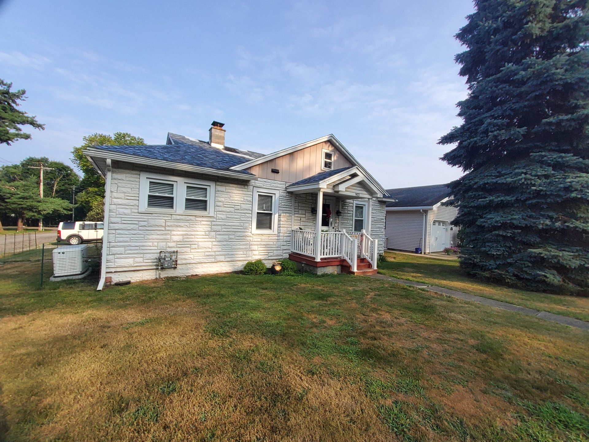 White house with peeling paint, overgrown lawn, and a large evergreen tree.